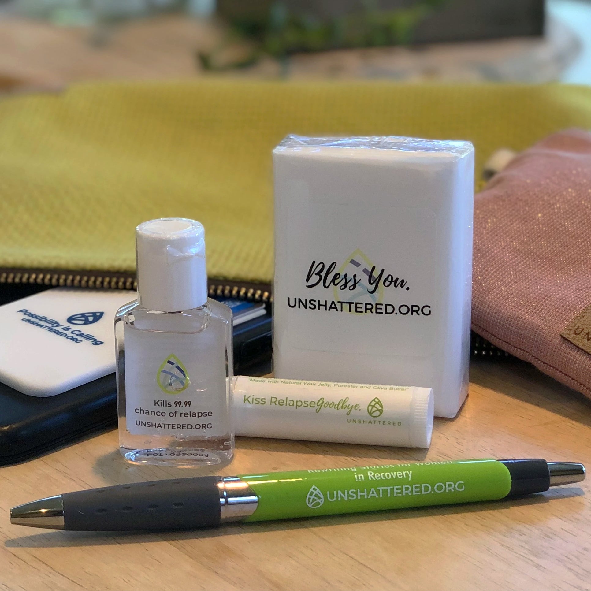 One green pen, one small bottle hand santizer, one tissue pack, one phone with rubber phone case attached to the back sit on a wood table peaking out of a small tan pouch.