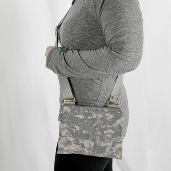 A woman wearing a gray long sleeve top with black jeans carrying a u.s army crossbody pouch with gray straps and gold hardware 