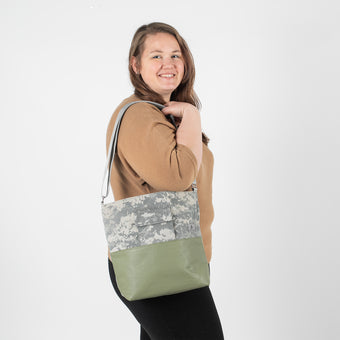 A woman wearing a tan top carrying an U.S Army tote with green luxury leather alternative on the bottom with a gray label and straps.