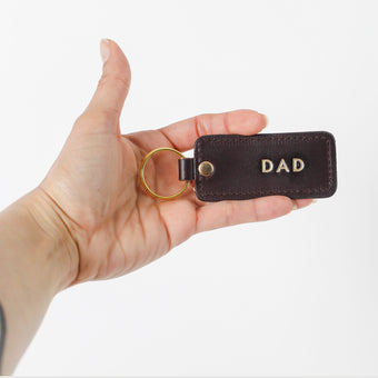 Hand holding a brown leather keychain with 'DAD' engraved on it against a white background
