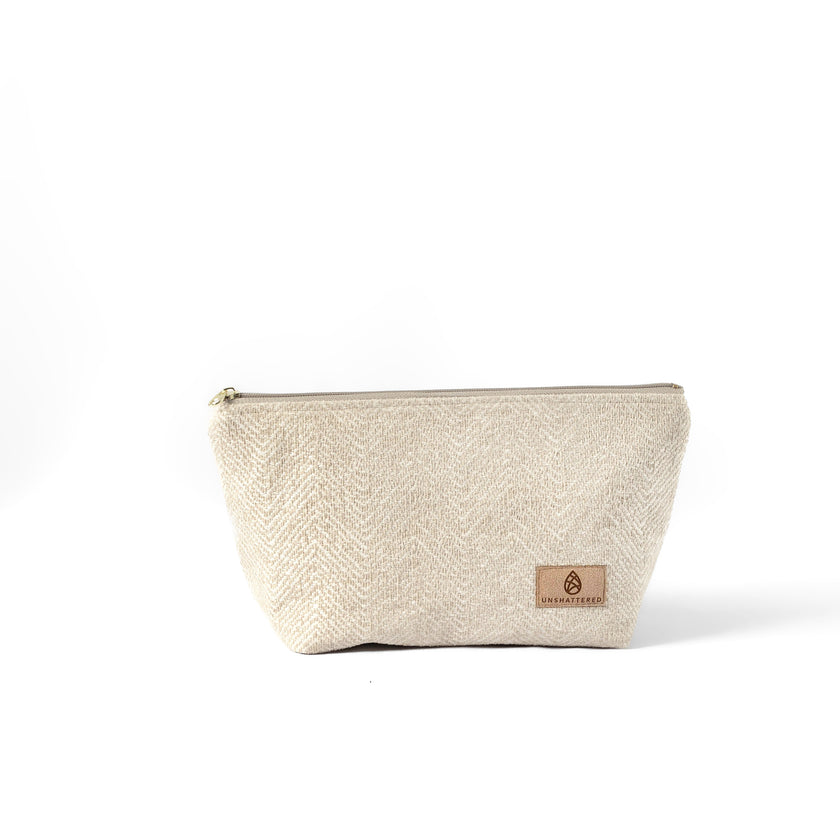 A beige, textured makeup bag named Theresa with a zip closure and a suede Unshattered label on a white background