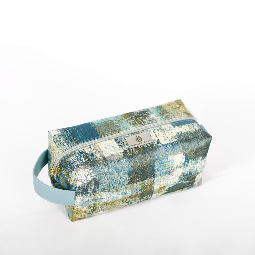 A toiletry bag made from upcycled fabric with a geometric pattern in shades of blue and green, featuring a zip closure and a light blue handle.