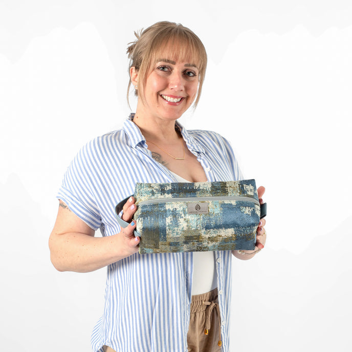 A woman carrying a toiletry bag made from upcycled fabric with a geometric pattern in shades of blue and green, featuring a zip closure and a light blue handle.