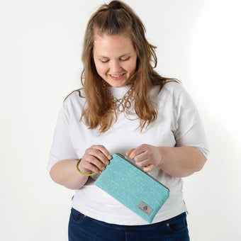 A woman opening a vibrant turquoise wristlet with blue dots, featuring a green wrist strap and gold-tone hardware named after Alicia.