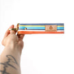 Hand holding a colorful striped key clip with 'Unshattered' branding on a white background named after Angel 