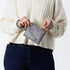 A woman opening a gray-blue clip pouch with a removable wristlet strap and gold-tone hardware, zippered closure, displayed against a white background named after Camille.