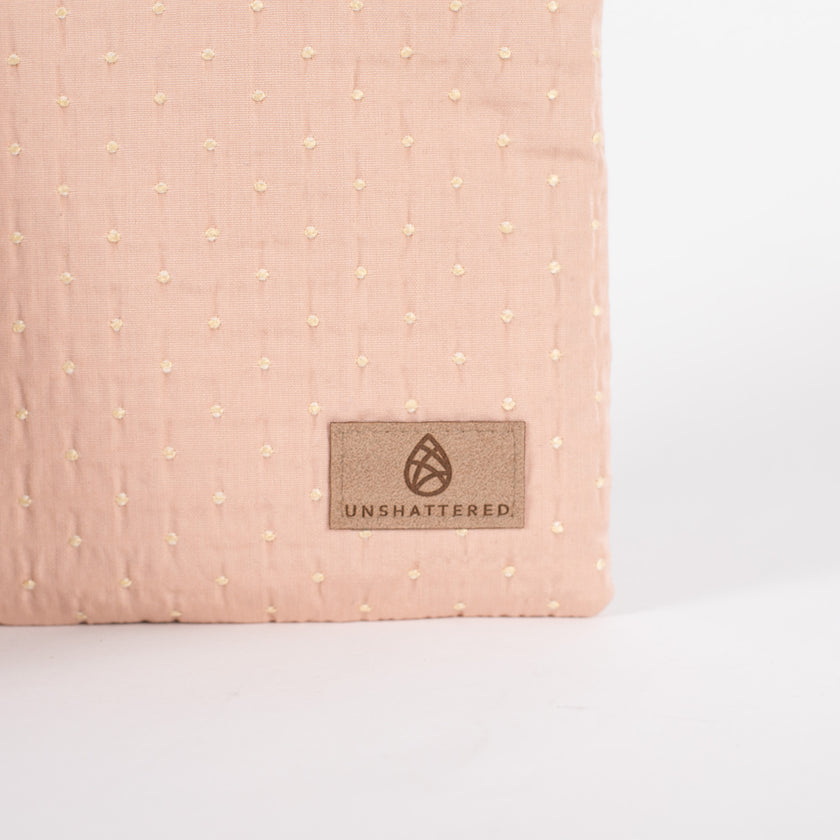 A pink and yellow dotted crossbody pouch featuring a tan-colored Unshattered label prominently displayed in the bottom right corner.