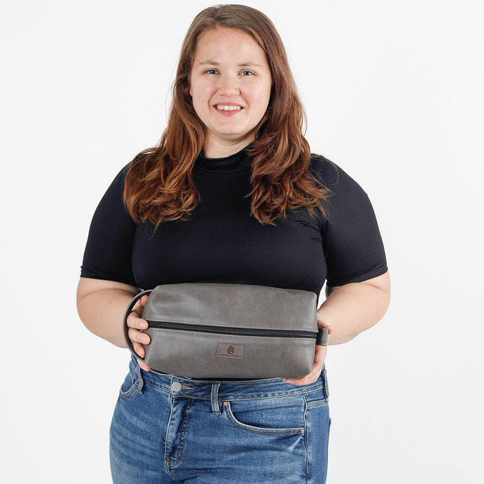A woman holding a gray leather toiletry bag with a zipper closure, a handle, and a brand label on the front named after Charles