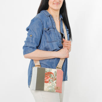 A woman carrying a crossbody pouch with a floral design on the front, featuring a mix of upcycled materials in multicolor, with a beige adjustable strap and gold-tone hardware named after Julie 