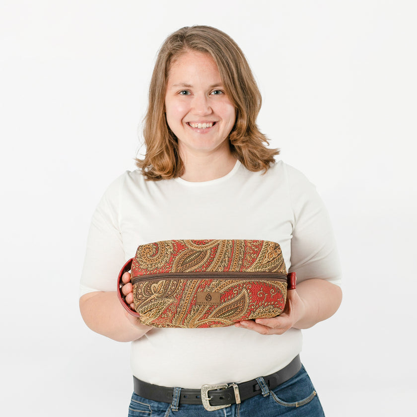 A woman holding a toiletry kit with a paisley pattern with reds, brown, green and gold named after Destiny
