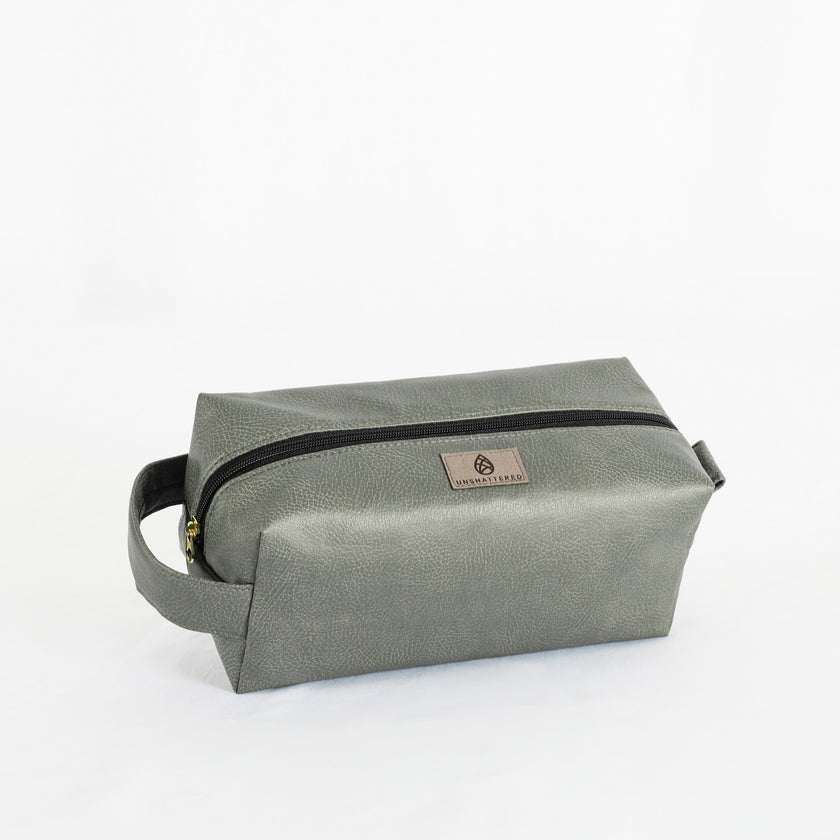 A gray toiletry kit with a zipper closure and a label displaying the brand name Unshattered named after Francine 