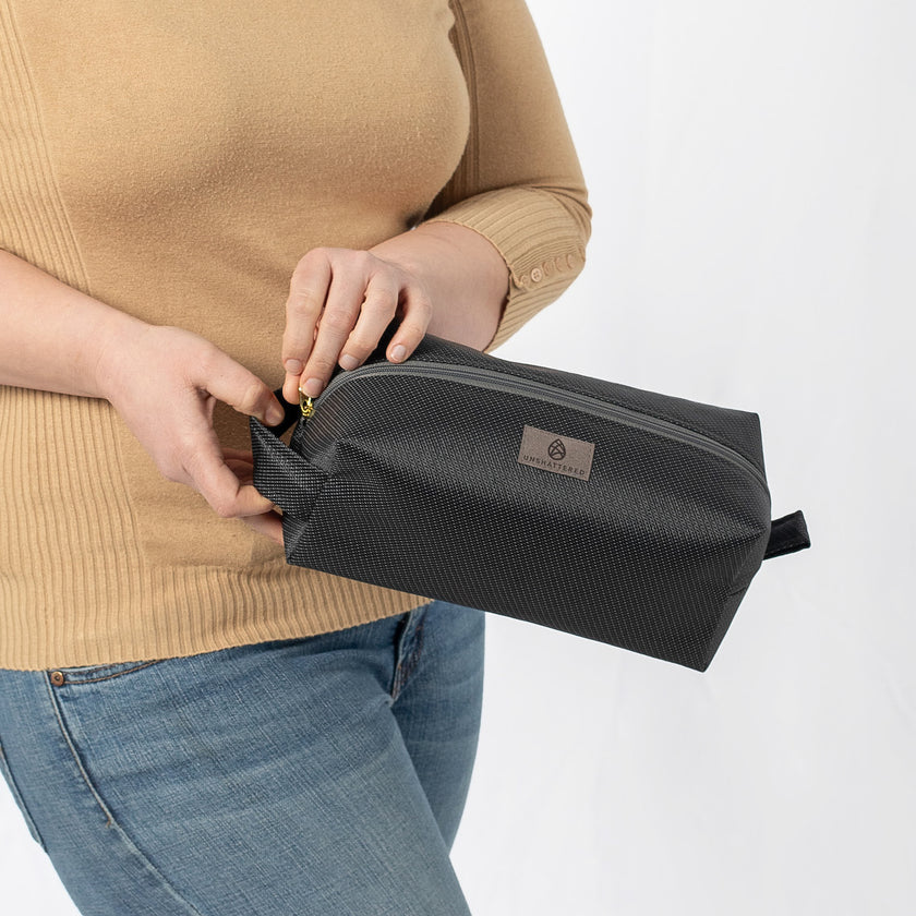 A woman opening a black toiletry kit with a gold toned zipper closure, made from outdoor furniture material  with a textured surface, featuring a gray Unshattered label 