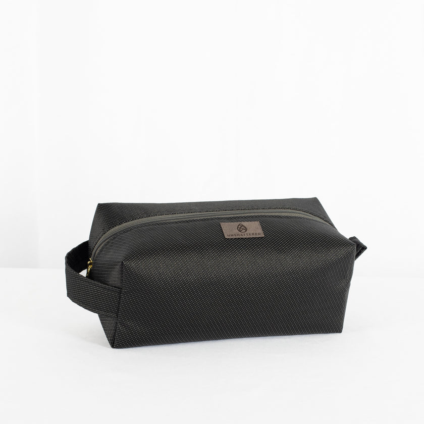 A black toiletry kit with a zipper closure, made from outdoor furniture material  with a textured surface, featuring a small brand tag attached to the zipper.