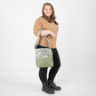 A woman wearing a tan top carrying an U.S Army tote with green luxury leather alternative on the bottom with a gray label and straps.