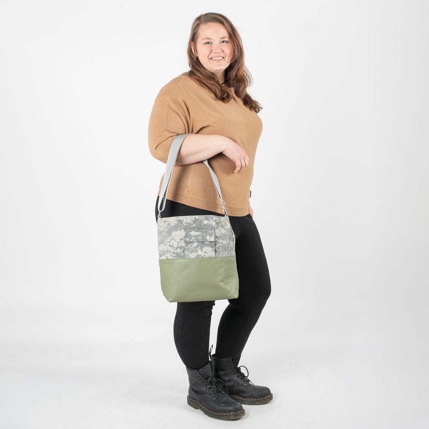 A woman wearing a tan top carrying an U.S Army tote with green luxury leather alternative on the bottom with a gray label and straps.