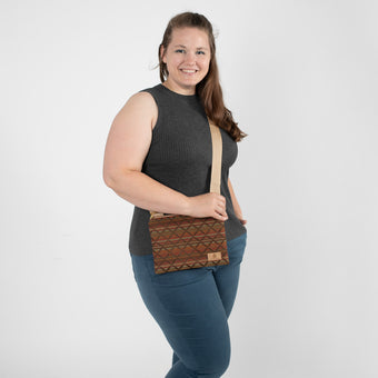 A woman smiling carrying a geometric-patterned fabric crossbody pouch, styled with a tan label, matching tan strap, and gold hardware.