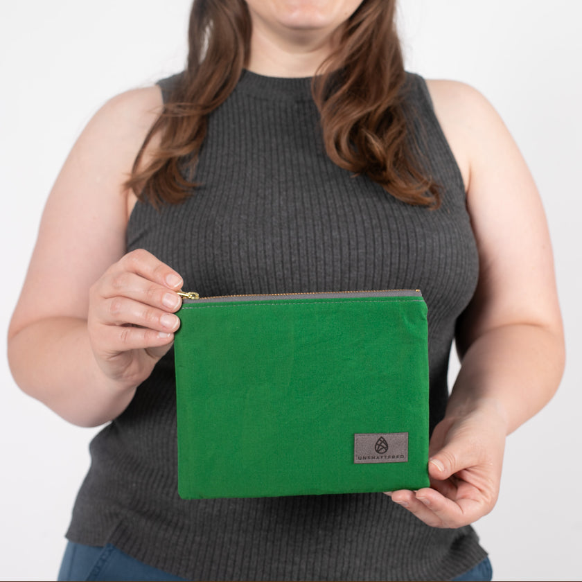 A woman  holding a textured green fabric zip pouch featuring a gray label and a matching gray zipper, shown in a detailed, up-close view to highlight the fabric quality.