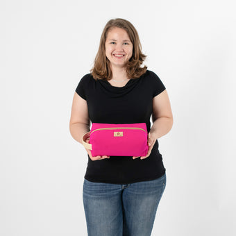 A woman wearing a black shirt with blue jeans holding a hot pink toiletry kit with cream accents and tan label 