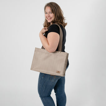 A woman wearing a black shirt with blue jeans smiling holding a tan zippered shopper tote bag with brown straps and gold hardware 