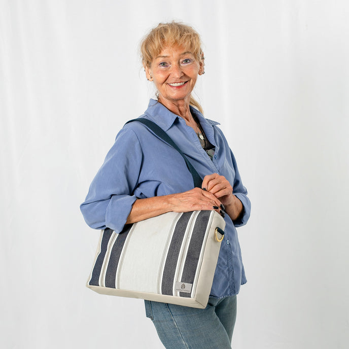A woman carrying a convertible handbag with a beige upcycled fabric, blue stripes, and navy-colored straps with gold-tone hardware.