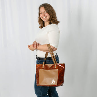 A woman wearing a white shirt with blue jeans holding a fall shiny brown unlined leather tote with a gold embossed logo in the bottom right