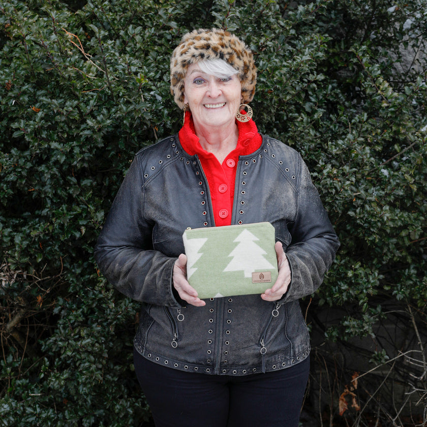 Woman holding a green zip pouch with a tree design against a green holly tree in the background