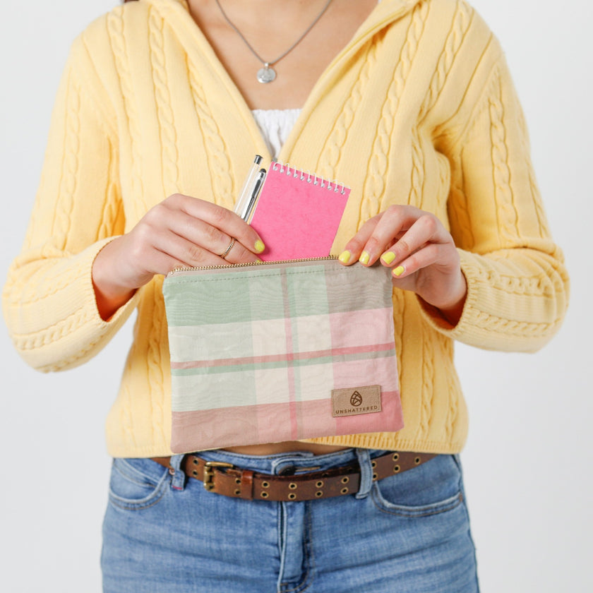 A woman holding a pink picnic plaid zip pouch putting items into the bag wearing a yellow sweater named after Kim 