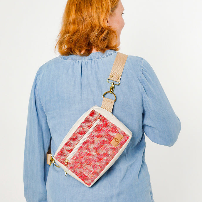 A woman wearing a pink/red  tweed errand bag with vegan leather accents, an adjustable strap, and gold-tone hardware. The bag features a front zip pocket, names after Korin 