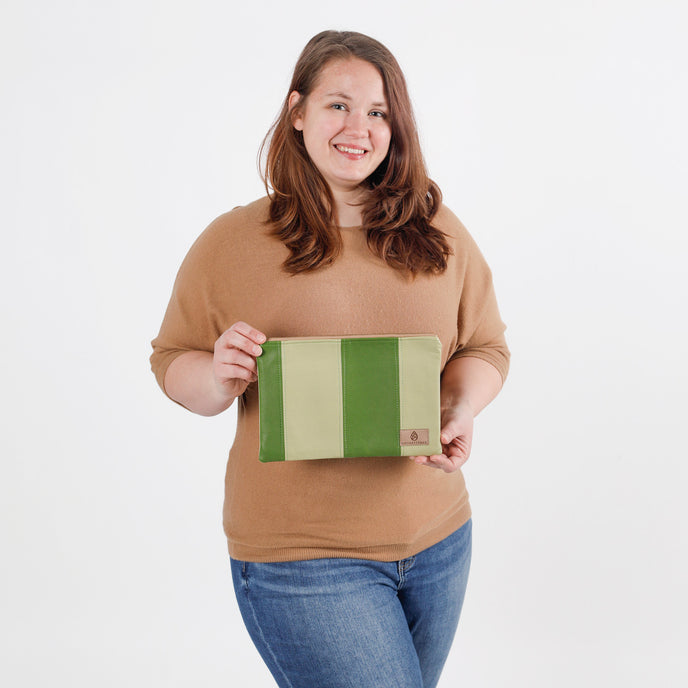 A woman wearing a tan sweater and blue jeans holding a multi-panel green large zip pouch against a white background