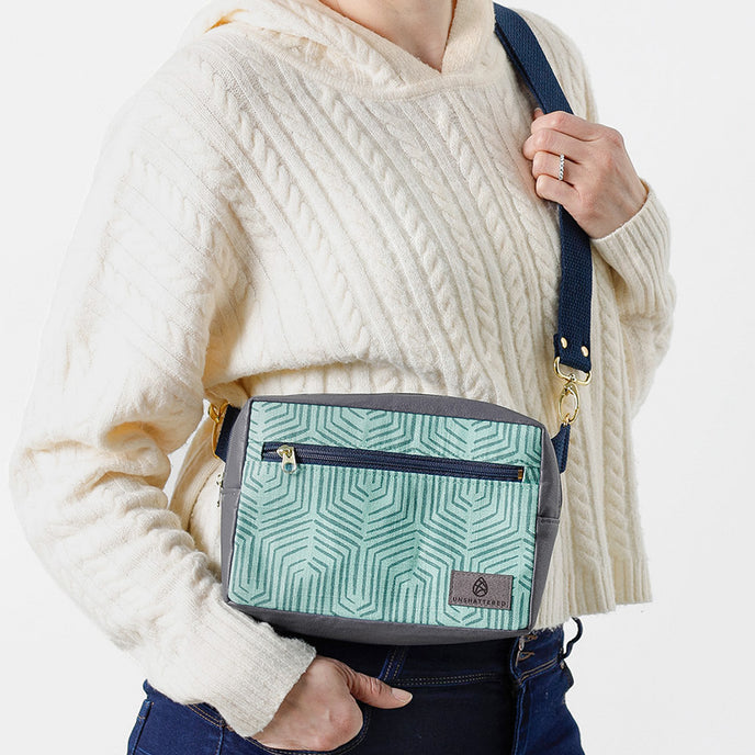 A woman carrying a small, blue geometric patterned handbag with an adjustable strap, front zip pocket, and gold-tone hardware, against a white background named after Kristine 