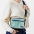 A woman carrying a small, blue geometric patterned handbag with an adjustable strap, front zip pocket, and gold-tone hardware, against a white background named after Kristine 