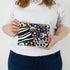 A woman holding a fun vibrant design zip pouch with a gray label and silver hardware named after Lella