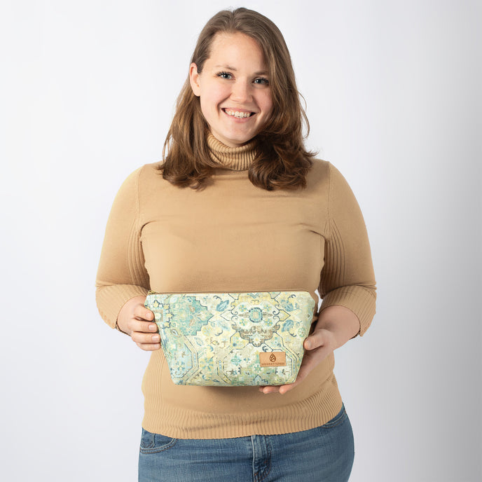 A woman smiling and holding this damask fabric makeup kit, featuring a tan brand label and a matching tan zipper.