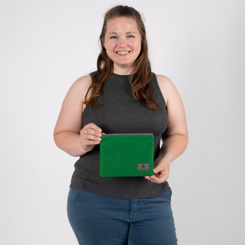 A woman smiling while holding a textured green fabric zip pouch featuring a gray label and a matching gray zipper.