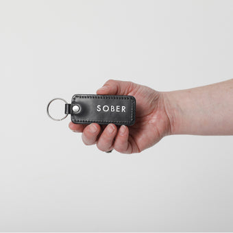 Hand holding a black leather key fob with 'SOBER' engraving on a white background