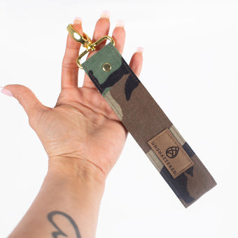 A woman holding a Woodland Camo key clip handcrafted from repurposed military uniform fabric, featuring a distinctive brown label and elegant gold-toned hardware in a lifestyle setting.