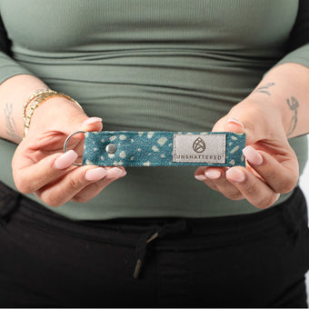 A person holding a teal circle fabric loop keychain with a gray label and silver tone hardware 