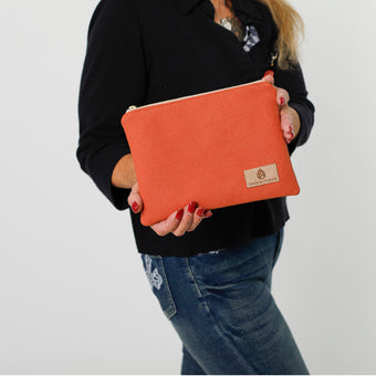A hand holding a orange pebble zip pouch bag with a tan Unshattered label, named after Elaina