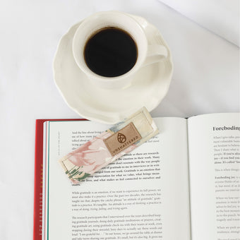 Cup of coffee on a saucer with an open book underneath, featuring an flower bookmark.