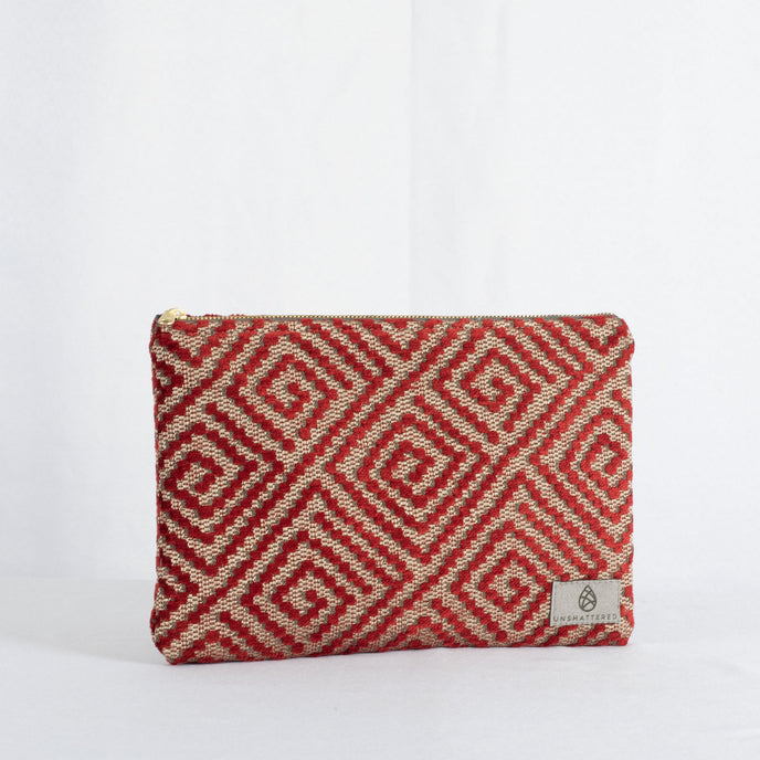 A large zip pouch with a geometric red and beige design and a gray fabric lining, featuring gold-tone hardware and a brand tag displaying 'Unshattered'.