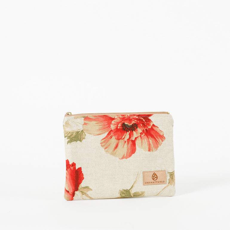 A zip pouch with a beige background and vibrant red floral accents, featuring gold-tone hardware and a brand tag displaying 'Unshattered' named after Mayra.
