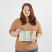 A woman holding a large zip pouch with striped velour fabric in beige, blue, and gray tones, featuring a gold-tone zipper named after Neenie.