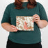 A woman holding a floral pattern zip pouch with a tan Unshattered label named after Reisha 