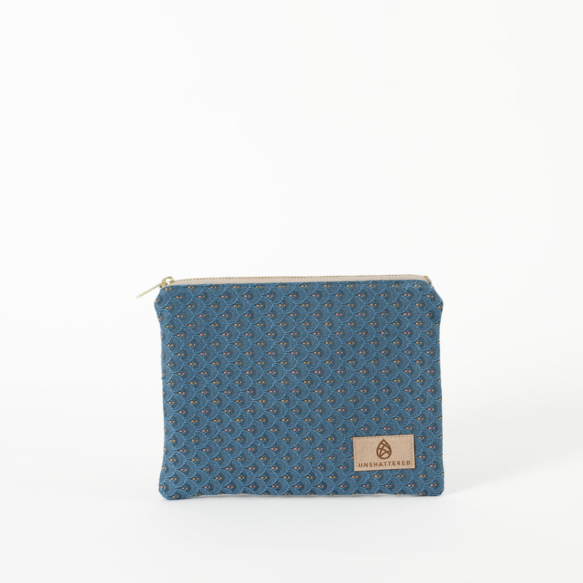 A small zip pouch with blue paisley fabric and gold-tone hardware, featuring a brand tag that reads Unshattered 