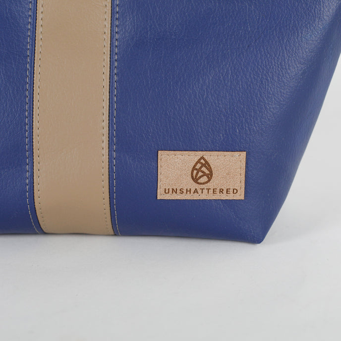 Blue and beige leather makeup bag with 'Unshattered' brand label on a light gray background