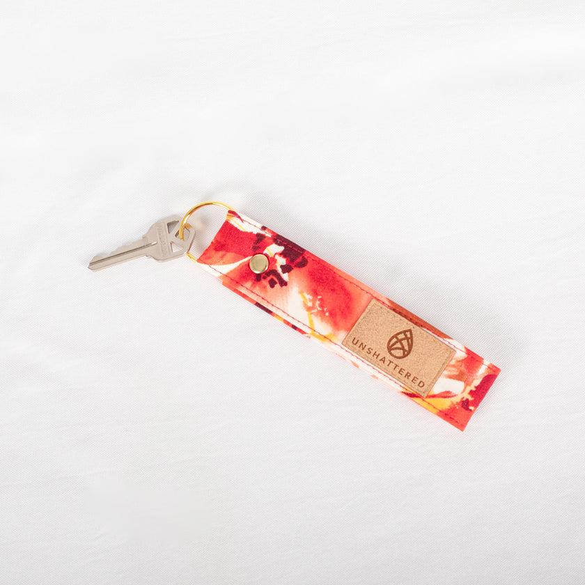 A gold-tone keyring attached to a red and orange floral patterned keychain with a label that reads Unshattered name after Susana.