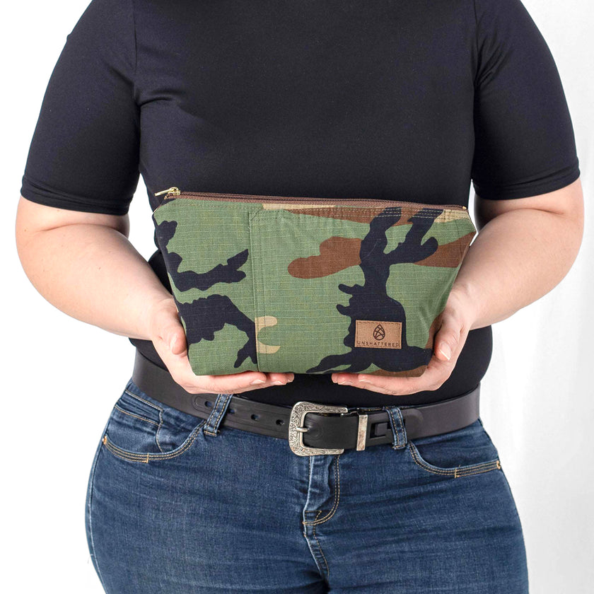 A woman holds a makeup bag handcrafted from upcycled woodland camouflage uniforms with a brown label and gold tone zipper