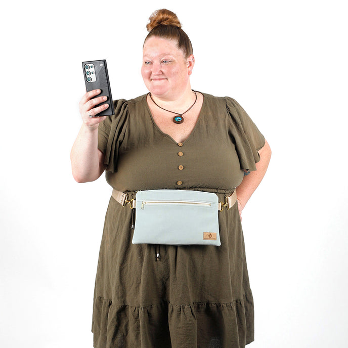 Woman looking at a phone while wearing a light blue 3-in-1 bag named George hands free belt bag style against a white background 