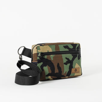 A horizontal crossbody errand bag with a woodland camo print, featuring a black adjustable strap, a front zip pocket, and an exterior drop-in pocket on the rear.