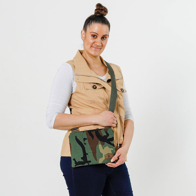 A woman wearing Woodland Camo Crossbody bag featuring a brown Unshattered label and green strap with gold hardware
